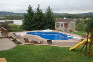 newly installed paver patio around a pool. a new poolhouse sits in the background next to three large evergreen trees