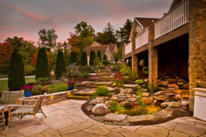 stone patio, wall steps, muclh beds, pond, and deck with landscape lighting mixed in