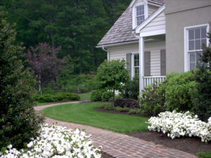 brick walkway around the side of a home. flower beds and grass surround the path with trees and shrubs