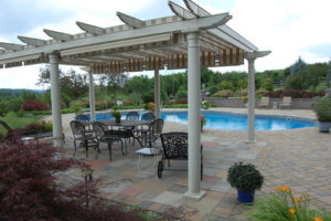 seating area underneath a pergola next to a pool. stone mulch used in flower beds stocked with a red japanese maple and other shrubs, flowers and more