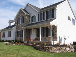 decorative stone wall and flower bed in front of a large home. fall pumpkins and decorations are mixed in on the porch and walk