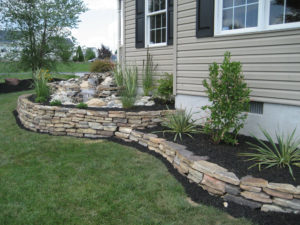 stone walls enclosing two flower beds on the side of the home. upper bed has water feature