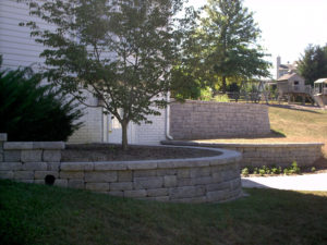 stone retaining wall in front of a raised flower bed with a tree and evergreens