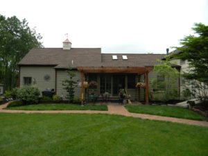 a pergola with a patio seating area beneath it. a brick walkway runs between two different doors and leads off behind the home.