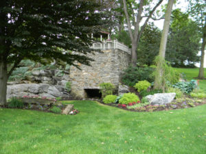 a large stone wall beneath a gazebo. several mulch beds with trees and shrubs surround the stone wall
