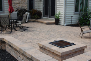 a paver patio, wall, and firepit behind a home. you can see outdoor seating to the left and right side