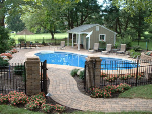 a paver walkway leads to a fenced in pool area with a pool house in the background