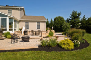 a paver patio area in back of a home. a small firepit and other outdoor furniture sit on the new patio. mulch beds surround the outer rim with various shrubs
