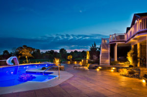 a pool, paver patio, and deck leading down to the pool. landscape lighting is mixed in throughout to light the backyard at dusk