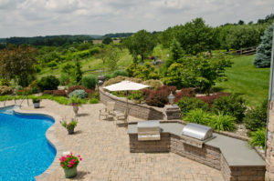 stone wall with built in grills, well maintained flower beds, and hardscaped patio area next to a pool