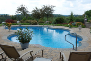 pool with nice hardscaped pavers around it. potted plants and outdoor furniture sit around the pool and nice landscaping is in the background