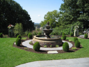 a circular fountain surrounded by several boxwoods in a mulch bed